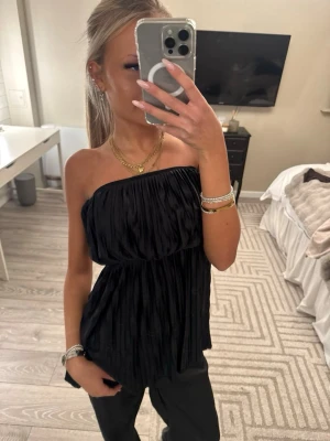 Svart plisserad tubtopp - Säljer en svart plisserad tubtopp med bandeau-modell. Toppen är axelbandslös och har ett snyggt fall med mycket volym. Perfekt att styla med smycken och vida byxor för en trendig look.