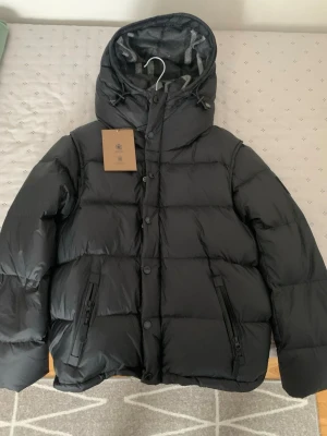 HELT NY Svart pufferjacka från Burberry - Säljer en svart pufferjacka från Burberry med huva och dragkedja samt knappar framtill. Jackan har två snedställda fickor med dragkedja och Burberry-logga på bröstet. Insidan av huvan har ett gråmelerat mönster. Perfekt för kalla dagar! PRIS KAN DISKUTERAS 