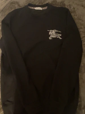 Svart sweatshirt från Burberry med tryck - Säljer en svart sweatshirt från Burberry i storlek M. Tröjan har ett stort vitt tryck av en riddare på häst på ryggen och ett mindre tryck på bröstet. Tillverkad i mjuk bomull och har klassisk rund hals och långa ärmar. Perfekt för dig som gillar stilrena designer med en twist.