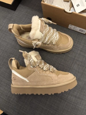 UGG lowmel - Chunky sneakers från UGG i beige mocka och mesh med fluffigt fårfoder på insidan. Skorna har breda, mönstrade snören i beige och vitt samt en rejäl platt sula. UGG-logga på sidan och bak. Perfekta för dig som vill ha både stil och komfort.