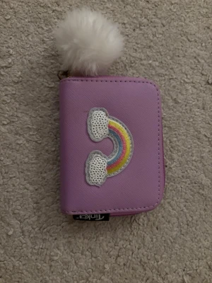 Lila plånbok med regnbågsdetalj - Söt lila plånbok från Tinks med broderad regnbåge och vita paljettmoln på framsidan. Plånboken har dragkedja runtom och en fluffig vit boll som nyckelring. Insidan har flera fack och ett myntfack med dragkedja. Perfekt storlek för kort och kontanter.
