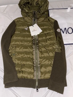 Grön overshirt från Moncler - Säljer en olivgrön pufferjacka från Moncler med stickade ärmar och huva. Jackan har dragkedja framtill, ribbade muddar och Moncler-logga på ärmen. Materialmix med polyester och stickad ull ger en schysst kontrast. Perfekt för kyliga dagar.
