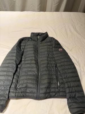 Mörkgrå pufferjacka från Colmar - Säljer en mörkgrå pufferjacka från Colmar med diskret logga på armen. Jackan har hög krage, två dragkedjeförsedda fickor och quiltad design. Insidan är ljusare 