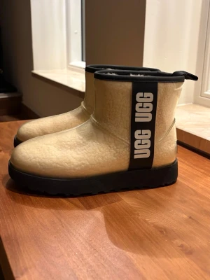 Beige vattentäta uggs  - Säljer ett par beige UGG boots med svart sula och  UGG-logga på sidan. Skorna har en genomskinlig yta över det fluffiga materialet och är vatten/snö täta, perfekta för höst och vinter💙 Storlek 36 och bra skick!