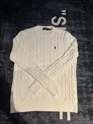 Vit kabelstickad tröja Polo Ralph Lauren - Vit kabelstickad tröja från Polo Ralph Lauren med klassisk rund halsringning och broderad marinblå logga på bröstet. Tröjan är långärmad och har ribbade muddar vid ärmslut och nederkant. Perfekt för lager-på-lager under hösten och vintern.