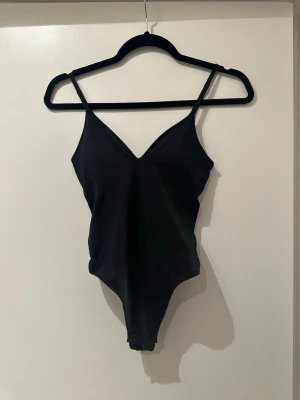 Svart body med smala axelband H&M - Svart body från H&M med smala justerbara axelband och v-ringad hals. Tillverkad i mjukt material med stretch för skön passform. Knäppning i grenen för enkel på- och avtagning. Enkel och stilren design som funkar till mycket.