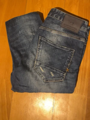 Blå slitna jeans från Scotch & Soda - Säljer ett par blå jeans från Scotch & Soda med snygga slitningar och distressed detaljer framtill. Jeansen har klassisk femficksdesign, normal passform och är tillverkade i jeansmaterial. Hör av er vid funderingar✌️