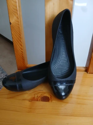 Svarta ballerinaskor från Crocs - Snygga svarta ballerinaskor från Crocs i syntetmaterial. Skorna har rund tå och en glansig detalj framtill som ger en stilren look. Perfekta för dig som vill ha bekväma och lätta skor till vardagen. Klassisk design med diskret Crocs-logga på sidan.