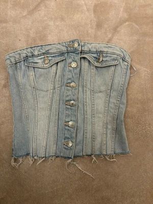 Ljusblå jeanskorsett från Zara - Säljer en ljusblå jeanskorsett från Zara med snygga silverfärgade knappar framtill och två dekorativa bröstfickor. Toppen är axelbandslös, har råa kanter nertill och smockad rygg för extra komfort.