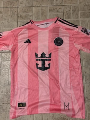 Rosa Inter Miami Messi matchtröja - Säljer en rosa Inter Miami CF matchtröja från Adidas med Messi 10 på ryggen. Tröjan har svarta detaljer, ränder och klubbmärke på bröstet samt MLS-logga och Apple TV-tryck på ärmen. Tillverkad i lätt polyester med texten 'Freedom to Dream' i nacken.
