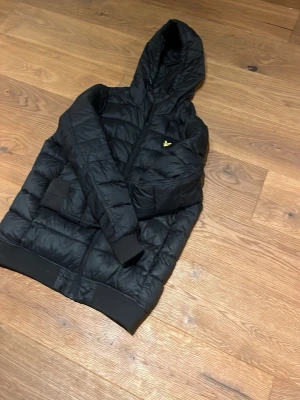 Svart pufferjacka från Lyle & Scott - Säljer en svart pufferjacka från Lyle & Scott med huva och gul logga på bröstet. Jackan är köpt för över 1000 kr och känns fortfarande ny.