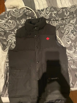 Svart dunväst från Canada Goose 2XS - Säljer en svart dunväst från Canada Goose i storlek 2XS. Västen har hög krage, stora fickor framtill och stängs med tryckknappar. Klassisk röd lönnlöv-detalj på bröstet och diskret logga på fickan. Materialet är polyester och bomull med dunfyllning.