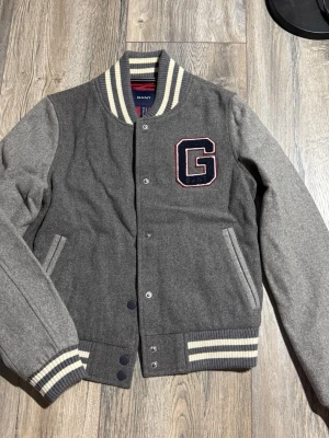 Grå bomberjacka från Gant - Snygg grå bomberjacka från Gant med stora G-broderiet på bröstet. Jackan har ribbade muddar och krage med vita ränder, tryckknappar framtill och två fickor.