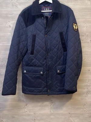 Morris quiltad jacka marinblå M - Quiltad marinblå jacka från Morris med snygga detaljer som emblem på ärmen, fickor med tryckknappar och bred krage i manchester. Fodret har prickigt mönster och jackan stängs med både dragkedja och knappar. Perfekt för höst och vinter.