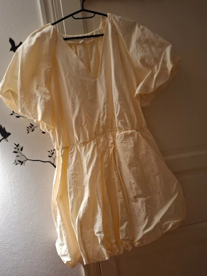 Ljus kortklänning med puffärm - Supersöt ljus kortklänning med puffiga kortärmar och v-ringning. Klänningen har en ballongformad kjol och resår i midjan för en avslappnad men ändå trendig look. Perfekt för sommarens alla tillfällen och enkel att styla med sneakers eller sandaler.