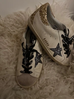 Golden Goose sneakers med glitter - Golden Goose sneakers i vitt med svart glitter  på sidan och guldglittrig häl. Svarta skosnören med logga och beige detalj på tån. Klassisk låg modell med platt sula och coola detaljer som verkligen sticker ut. Jag köpte dessa på en lopppas hyfsat nya men jag själv använt de fåtal gånger så de är i ganska bra skick. Jag upplever och tror att de är äkta så de får man avgöra själv eftersom att jag inte köpt från golden goose hemsida!! Otroligt snygg och gör hel ens outfit!!🥰pris går att diskutera!