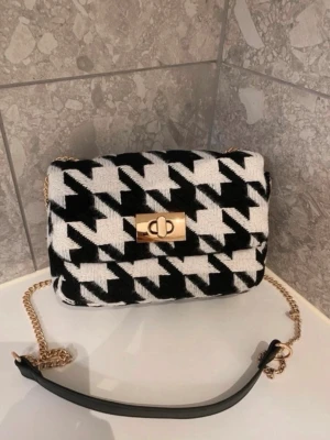Liten axelremsväska - Liten axelremsväska från H&M i säljes. Väskan är helt oanvänd och prislappen sitter kvar.  Är i svart/hundtandsmönstrad.   Perfekt till hösten/vintern!