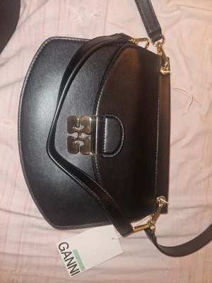 Svart axelväska från Ganni i skinn - Snygg svart axelväska från Ganni i slätt skinn med guldfärgade metalldetaljer och präglad logga på både rem och insida. Väskan har en unik formad frontflik och justerbar axelrem. Insidan är fodrad i beige mockaimitation och har en innerficka.