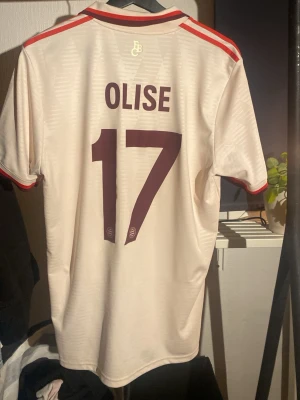Äkta Adidas Bayern München Olise tröja M - Säljer en ÄKTA beige Bayern München matchtröja från Adidas med Olise och nummer 17 på ryggen. Tröjan har röda detaljer på krage och ärmslut, samt klubbmärke och Adidas-logga på bröstet. Slim fit-modell i lätt och ventilerande material.