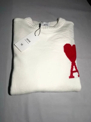 Ami Paris herr - Vit Ami Paris med rött hjärta Stilren och tidlös vit Ami Paris-tröja prydd med det ikoniska röda hjärtat – en perfekt kombination av fransk elegans och modern streetwear. Tröjan har en minimalistisk design som gör den lätt att matcha till både vardagliga och mer uppklädda outfits. Tillverkad i mjukt och bekvämt material för hög komfort hela dagen. Ett måste i garderoben för dig som uppskattar exklusiv design med en diskret men tydlig signatur.