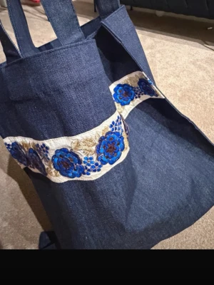 Blå shoppingväska med blommigt band - Unik shoppingväska i mörkblå denim med breda axelband och ett dekorativt band med broderade blå blommor och gulddetaljer. Perfekt storlek för att bära böcker, laptop eller annat du behöver under dagen. Väskan har en enkel, stilren form och känns både trendig och personlig.