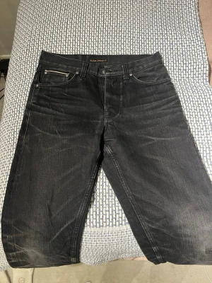Svarta Nudie Jeans, W29 L30 - Svarta jeans från Nudie Jeans med klassisk femficksdesign och subtila slitningar. Jeansen har en rak passform och är tillverkade i slitstark bomull. Snygga kontrastsömmar och Nudie Jeans-logga på bakfickan ger extra stilpoäng. Detta är också salvage som ger det lite dyrare priset