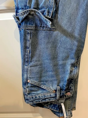 Blå cargojeans med raka ben - Säljer ett par blå cargojeans med raka ben och stora fickor på sidorna. Jeansen har klassisk femficksdesign, bälteshällor och stängs med knapp och dragkedja. Perfekta för dig som gillar streetstyle och vill ha extra plats för prylar.