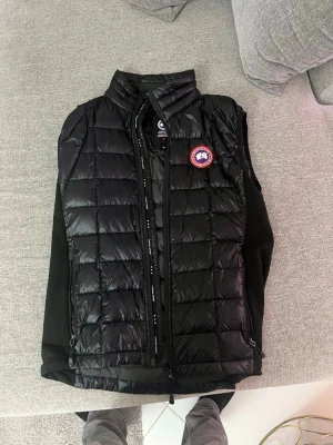 Svart dunväst från Canada Goose - Säljer en svart dunväst från Canada Goose med klassisk quiltad design och hög krage. Västen har dragkedja framtill och den ikoniska Canada Goose-loggan på bröstet. Perfekt att slänga över en hoodie eller tröja för extra värme.