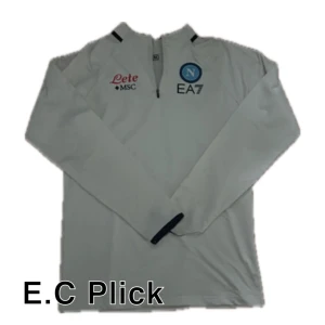 Vit långärmad Napoli EA7 fotbollströja - Snygg vit långärmad fotbollströja från Napoli med halv dragkedja, klubbmärke och sponsorloggor på bröstet. Tillverkad i ett lätt och funktionellt material som passar perfekt för träning eller match. Stilren design med svarta detaljer vid krage och ärmslut.