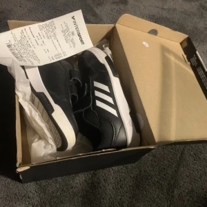 Svarta Adidas sneakers med kardborre - Snygga svarta Adidas sneakers med vita detaljer och klassiska tre ränder på sidan. Skorna har kardborreband för enkel påtagning och en vit, platt sula med svart mönster. Ovandelen är i syntetmaterial med textur och logga på hälen och sulan.