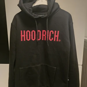 Svart HOODRICH hoodie med röd logga - Svart hoodie från HOODRICH i storlek small. Stor röd logga framtill, klassisk känguruficka och snörning i huvan. Perfekt för dig som gillar streetwear och vill sticka ut med en fet logga. Mjuk och skön bomullskänsla.