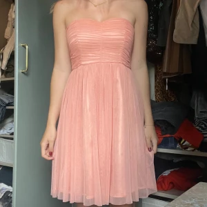 Ljusrosa axelbandslös klänning - Superfin ljusrosa axelbandslös klänning med plisserad överdel och vippig kjol. Klänningen har dragkedja i ryggen och är sydd i ett tunt, flowy material som ger en romantisk känsla. Perfekt för dig som vill ha en söt och stilren look.