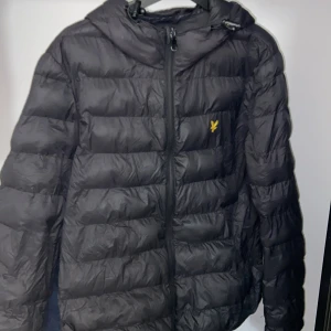 Svart dunjacka från Lyle & Scott - Svart dunjacka från Lyle & Scott med huva och gul logga på bröstet. Jackan har quiltad design, dragkedja framtill och justerbar snörning vid huvan. Perfekt för kalla dagar och har en modern, sportig look.