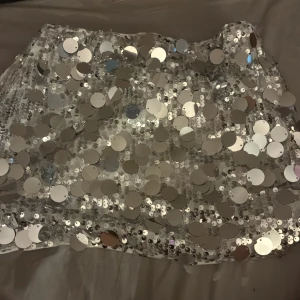 Glittrig silverkort kjol med paljetter - Supersnygg kort kjol täckt av stora silvriga paljetter som verkligen fångar ljuset. Kjolen har vit bas och elastisk midja för skön passform. Perfekt för dig som vill sticka ut och glänsa! Fodrad insida för extra komfort.
