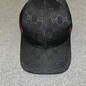 Svart keps med gg - Snygg svart keps frånmed det klassiska -monogrammet över hela kepsen. Kepsen har en böjd skärm och detaljer i rött och grönt på sidorna. Tillverkad i slitstark bomull och har justerbar rem baktill för perfekt passform.