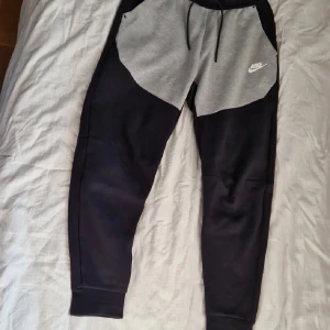 Nike mjukisbyxor i svart och grå - Snygga mjukisbyxor från Nike med svart och ljusgrå färgblock, klassisk logga på benet och snörning i midjan. Perfekta för chill eller träning, med skön passform och mjukt material. Elastiska muddar vid benslut och praktiska fickor på sidorna.