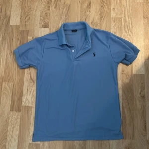 Blå pikétröja från Polo Ralph Lauren - Snygg blå pikétröja från Polo Ralph Lauren med klassisk krage och två knappar. Tröjan har broderad logga på bröstet och är tillverkad i bomull. Perfekt för dig som gillar stilren och tidlös design.