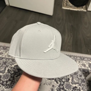 Ljusgrå Jordan keps snapback S/M - Snygg ljusgrå snapback-keps från Jordan med broderad Jumpman-logga framtill. Kepsen har rak skärm, justerbar snapback baktill och är tillverkad i polyester. Perfekt för dig som vill ha en clean och sportig stil.  Är helt oanvänd 