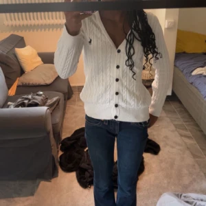 Vit kofta från Ralph Lauren - Vit kabelstickad kofta från Ralph Lauren med svarta knappar och broderad logga på bröstet. V-ringad modell med lång ärm och ribbade muddar. Perfekt att slänga över en t-shirt eller linne för en klassisk look. Pris kan diskuteras 