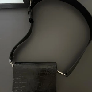 Svart axelväska i croco-mönster - Säljer en svart axelväska med croco-mönstrad yta och bred axelrem. Väskan har en stilren, rektangulär form och silverfärgade metalldetaljer.