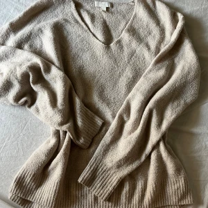 Beige stickad v-ringad tröja - Mysig beige stickad tröja med v-ringning och långa ärmar. Tröjan har breda ribbade muddar vid ärmslut och nederkant, vilket ger en avslappnad och trendig look. Perfekt för lager-på-lager och chill dagar.