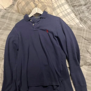 Marinblå långärmad pikétröja Polo Ralph Lauren - Snygg marinblå långärmad pikétröja från Polo Ralph Lauren med klassisk krage och knappar framtill. Ikoniska röda loggan broderad på bröstet. Perfekt för en stilren och avslappnad look.