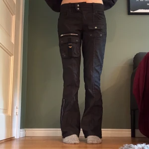 Svarta cargopants från BDG Urban Outfitters - Svarta, lågmidjade cargopants från BDG Urban Outfitters med flera fickor, dragkedjor vid bensluten och coola detaljer som spännen och nitar. Bootcut. Använda kanske 2 gånger.