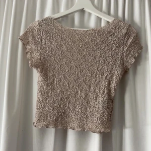 Beige spetsig croppad topp från H&M - Säljer en beige croppad topp i spets med korta ärmar och vågig kant nertill. Toppen har ett fint spetsmönster över hela och är lätt genomskinlig, perfekt att styla med jeans eller kjol. Sitter tight och har en feminin vibe.