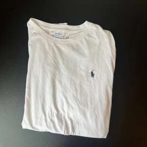 Vit t-shirt från Polo Ralph Lauren - Vit t-shirt från Polo Ralph Lauren i mjuk bomull. Klassisk passform med korta ärmar och rund halsringning. Ikoniska broderade loggan i mörkblått på bröstet. Perfekt basic-plagg med stilren look.