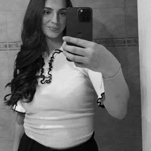 Vit ribbad blus med volang från Holly & Whyte - Supersöt vit ribbad blus från Holly & Whyte med svarta volangdetaljer vid krage, ärmslut och längs knäppningen. Blusen har trekvartsärm och pärlknappar framtill. 