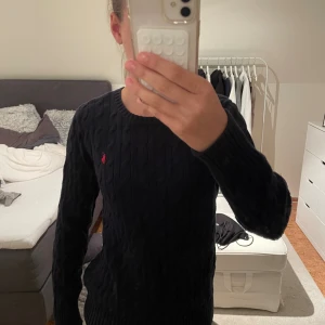 Mörkblå stickad tröja från Ralph Lauren - Kabelstickad mörkblå tröja från Polo Ralph Lauren med rund halsringning och röd broderad logga på bröstet. Tröjan har lång ärm och ribbade muddar vid ärmslut och nederkant. Perfekt för lager-på-lager och snygg till jeans. Skulle säga att den är mer i storlek M