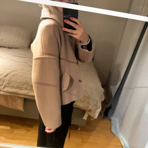 Beige kappa med krage och fickor - Säljer en beige kappa i kortare modell med klassisk krage och stora fickor framtill. Jackan har knappar och en loose passform som ger en chill vibe. Perfekt för lager-på-lager och snygg till både jeans och kostymbyxor.