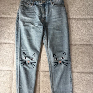 Ljusblå jeans med broderade kattansikten - Unika ljusblå jeans från Monki med raka ben och broderade kattansikten på knäna. Klassisk femficksmodell med hög midja och dragkedja. Jeansen är tillverkade i bomull och har en avslappnad, normal passform. Perfekta för dig som vill sticka ut med en lekfull detalj. Kortare modell, passar mig som är ca 160cm.