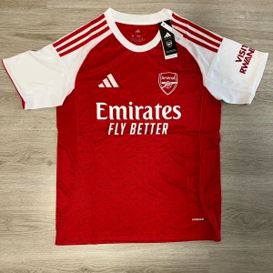 Arsenal hemmatröja Adidas röd/vit M - Snygg Arsenal hemmatröja från Adidas i rött och vitt med klubbmärke och sponsortryck. Tröjan har korta ärmar, klassiska Adidas-ränder på axlarna och diskret mönster i tyget. Tillverkad i lätt och ventilerande material (AEROREADY).
